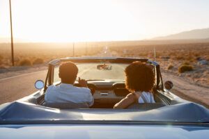 Rear View Of Couple On Road Trip Driving Classic Convertible Car Towards Sunset